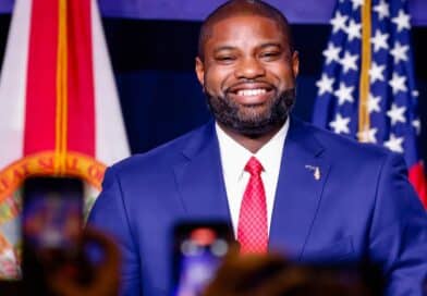Byron Donalds em evento político em Miami com bandeiras da Flórida e dos EUA